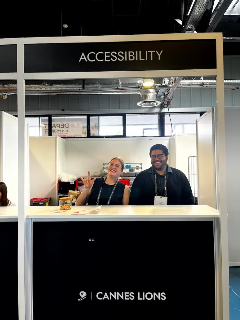 Two ICS team members stand behind the Cannes Lions accessibility desk, where attendees could request support. One of them holds up the ILY handshape under a sign reading 'Accessibility'.