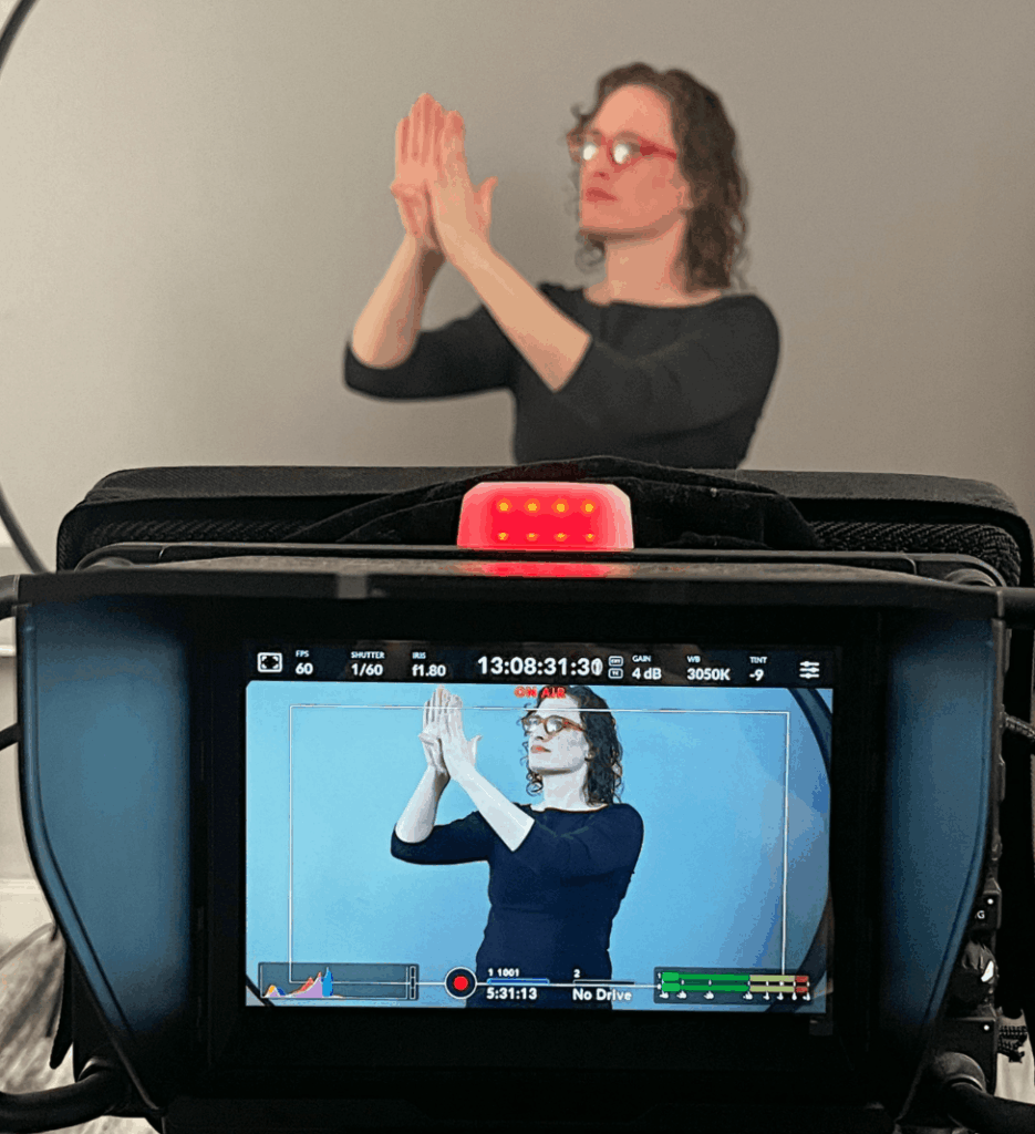 Over-the-shoulder view of the camera monitor mid-take, the Deaf interpreter's signing framed on the screen with the on-air indicator lit and exposure settings showing along the top.