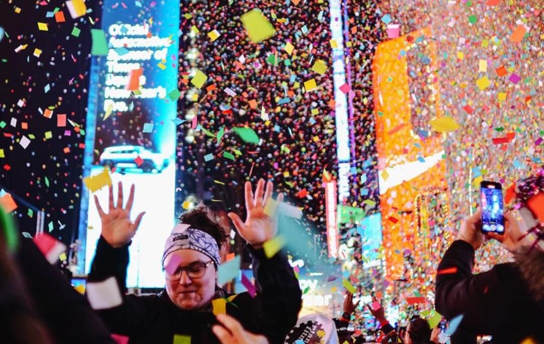 Confetti falling over the crowd in Times Square at midnight, an attendee in a headband in the foreground with both hands raised and a phone filming beside her.