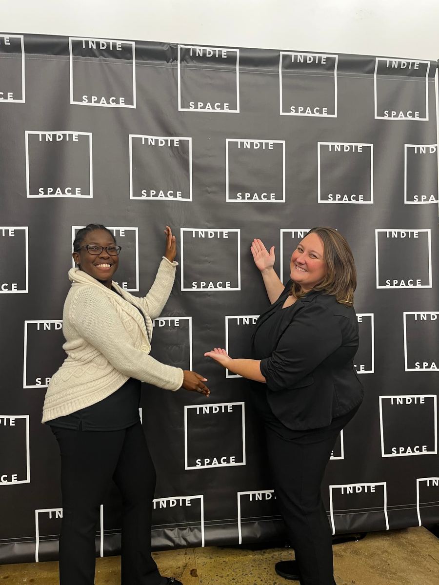 Two women with big smiles posing in front of a black and white banner with the words “Indie Space” on it. At the fundraiser for Indie Space’s theatre event.