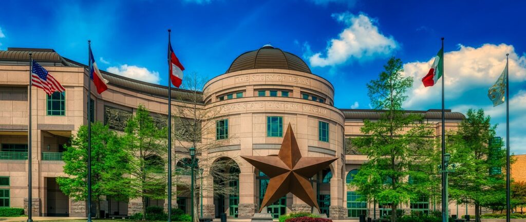 A domed stone museum rotunda lettered The Story of Texas above Texas State History Museum, a large bronze star standing on a plinth in the plaza in front of it, flagpoles on either side.
