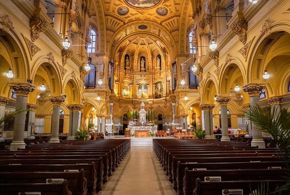 The nave of the Church of St. Francis Xavier looking toward the altar: gilded arches and Corinthian columns down both sides, painted panels above the sanctuary, and empty wooden pews either side of the centre aisle.