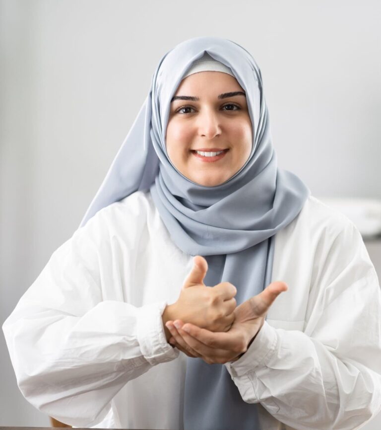 A smiling woman wearing a grey-blue hijab and white top; one hand is closed with the thumb pointing up, resting on the open palm of her other hand.