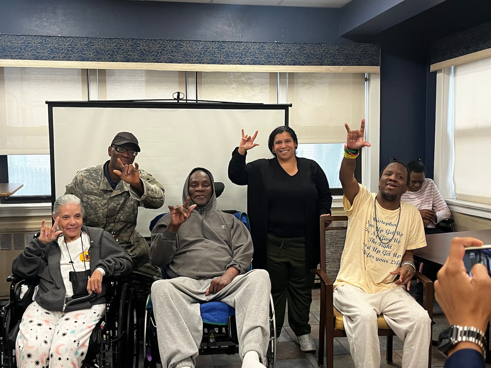 A group of people at the Bronx Senior Citizen Center holding one hand up with the sign ILY, meaning love.