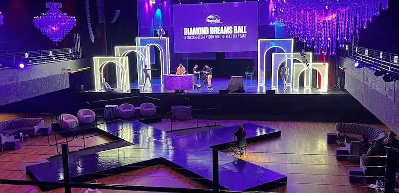 A wide view of the Diamond Dreams Ball room. Lit white arches frame a stage beneath a screen reading 'Diamond Dreams Ball: a crystal-clear vision for the next ten years', with a runway, lounge seating, and chandeliers on the floor below.
