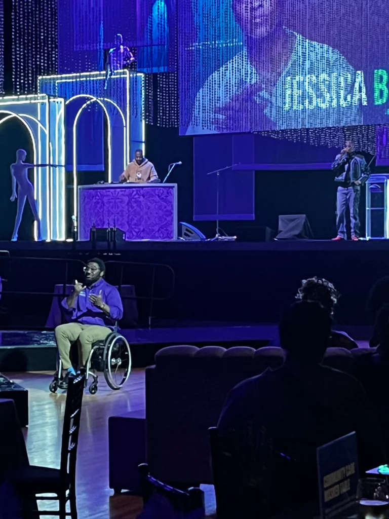 An ASL interpreter who uses a wheelchair signs from the floor in front of the Diamond Dreams Ball stage while a DJ and a performer work on the stage behind him.