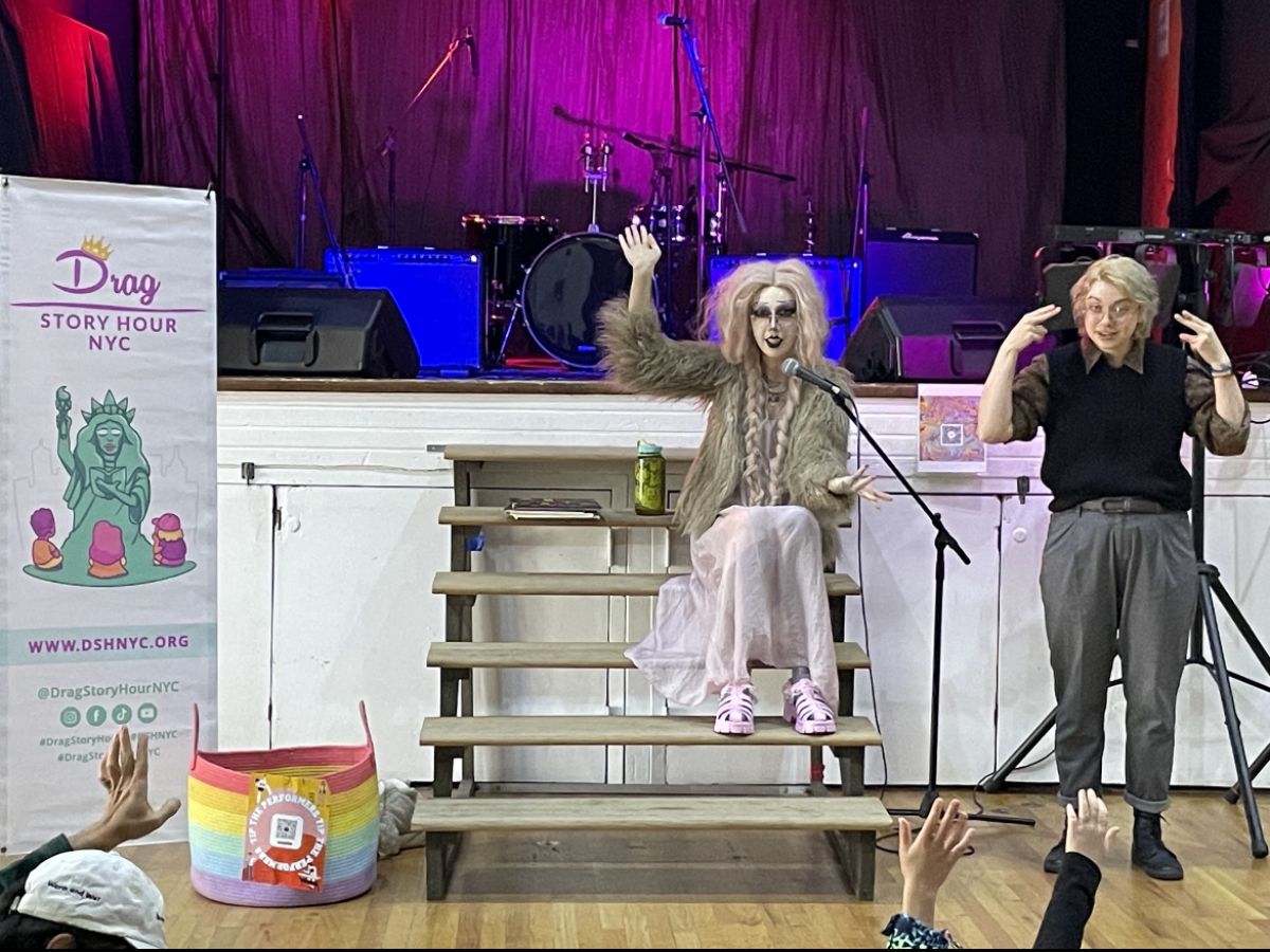 A queen sits on stairs in front of a stage with a drum set. To the queen’s left is the DSH banner, and to the right is an ASL interpreter.