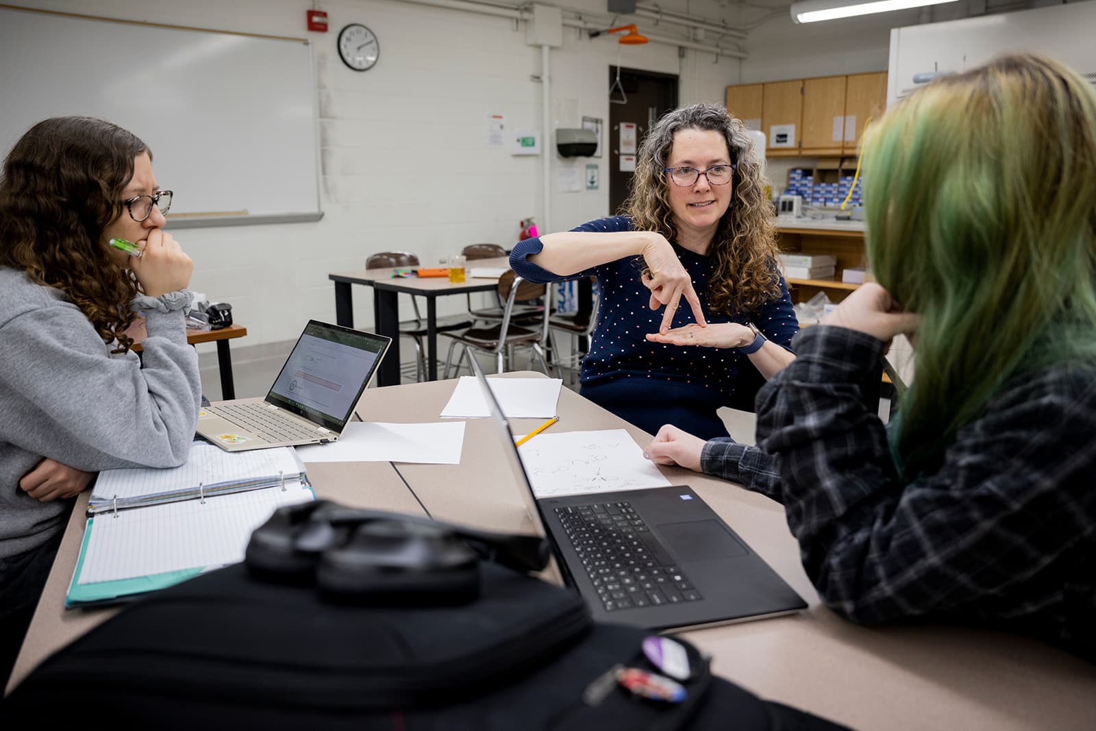 An interpreter signing across a classroom worktable to two students sitting with open laptops and handwritten notes.