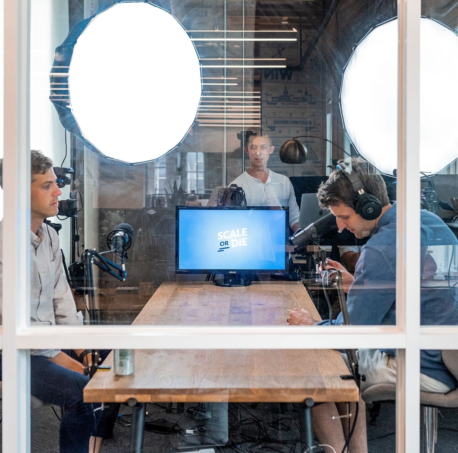 Three men record a podcast in a glass-walled studio with ring lights and monitors