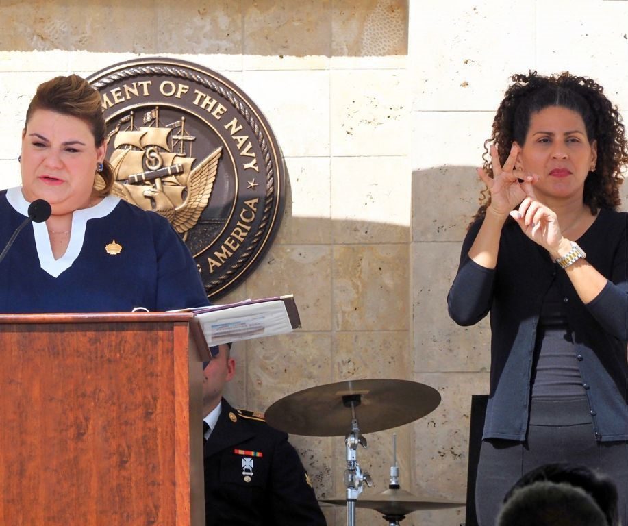An interpreter signs beside a speaker at a podium during an outdoor Navy ceremony