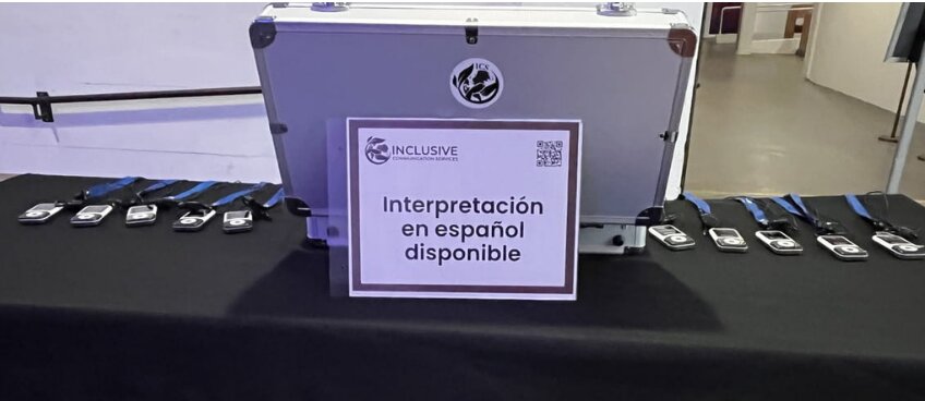 The distribution table at another venue: the ICS 'Interpretación en español disponible' sign in front of a closed flight case, with rows of receivers and lanyards laid out on the black cloth either side.