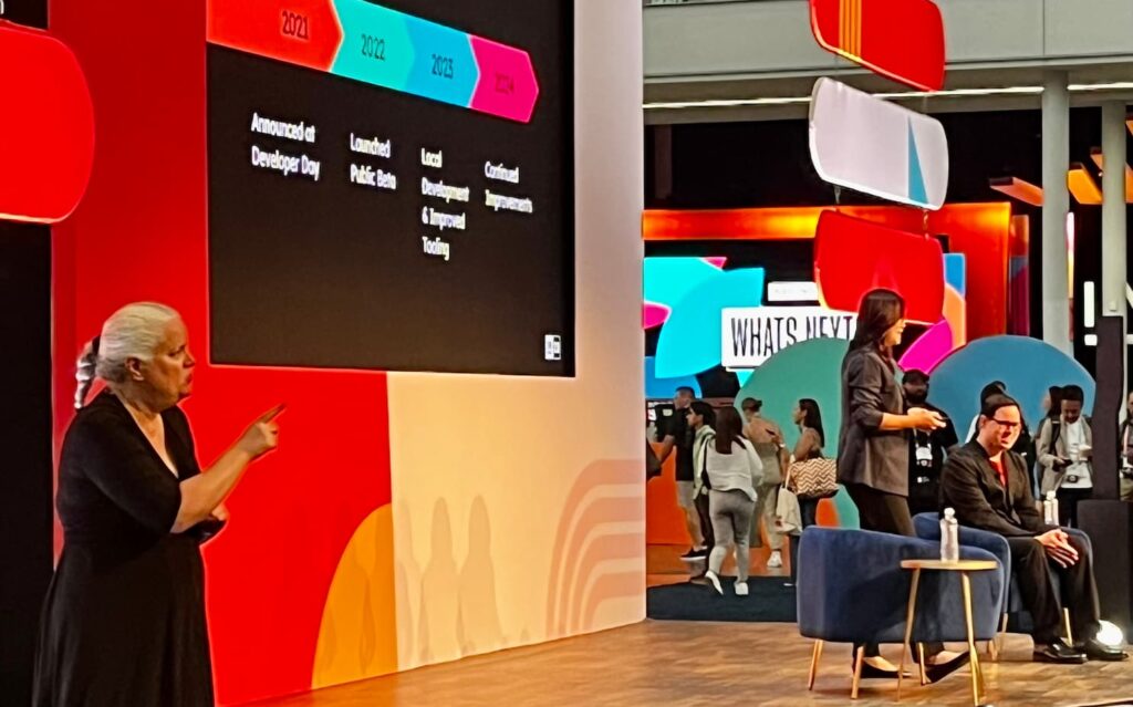An ASL interpreter signs beside the presentation screen in an open expo-floor session space, with a seated speaker and attendees walking past the brightly coloured INBOUND set behind her.