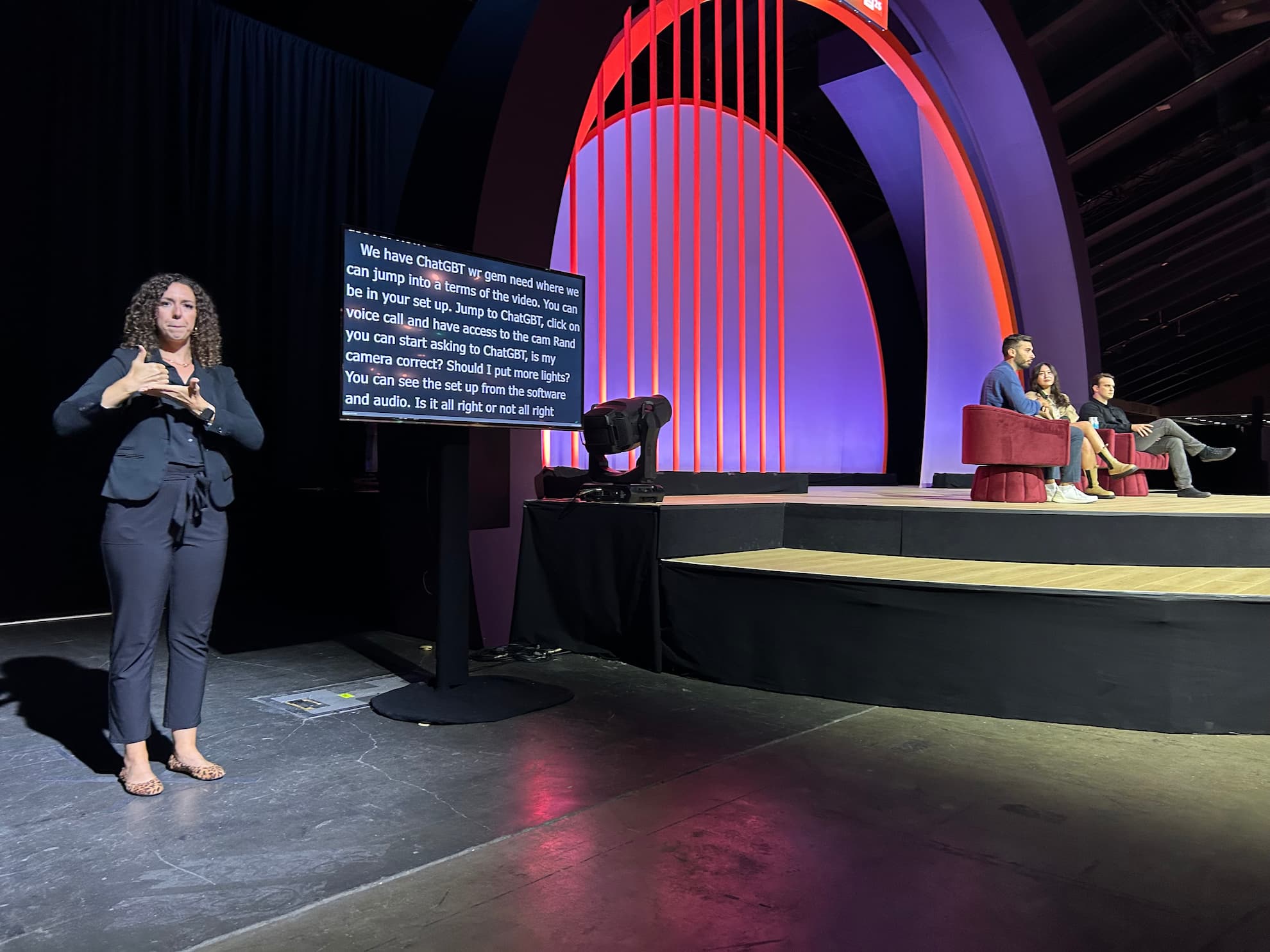 An ASL interpreter signs at floor level in front of a CART caption monitor while three panelists sit on the arched INBOUND stage to the right, the empty auditorium floor stretching between them.