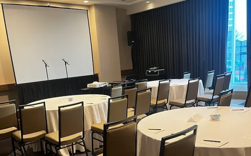 Before the event: an empty hotel meeting room set for the workshop, with two microphone stands in front of a projection screen and round tables laid out with handouts.