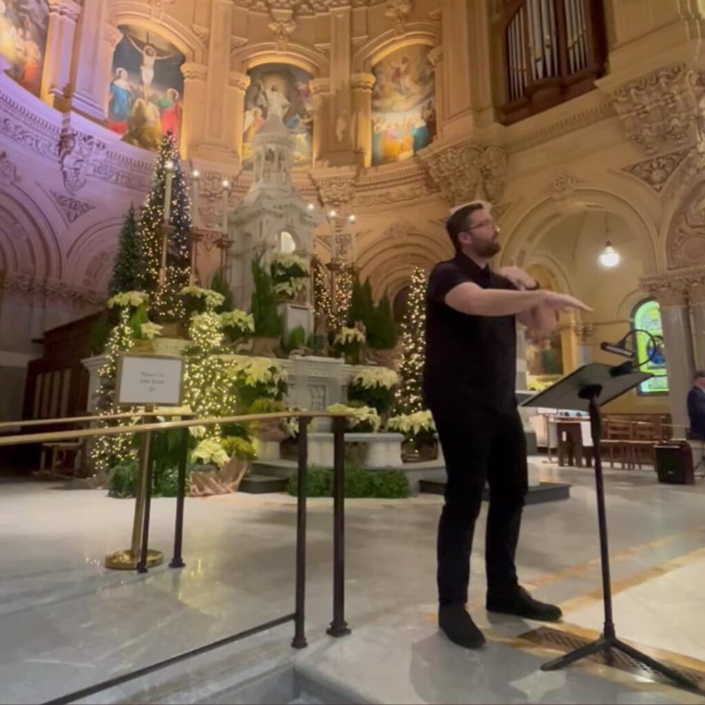 An ASL interpreter in black signs from the sanctuary steps during Christmas Mass, standing beside a music stand and microphone with lit Christmas trees, poinsettias, and the crèche behind him.