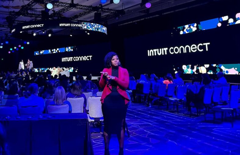 An ASL interpreter in a red blazer signs on the ballroom floor in front of the main stage, with 'Intuit Connect' on the screens above and rows of seated attendees lit in blue behind her.