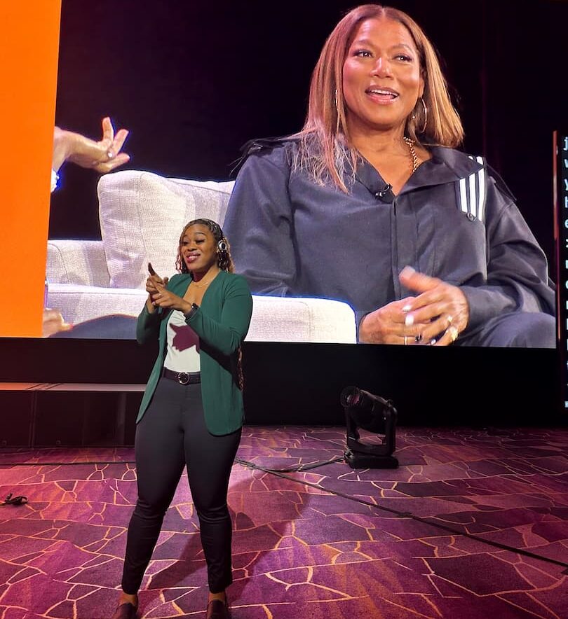 An ASL interpreter signs at the front of the stage while the keynote speaker she is interpreting appears on the screen behind her, seated in an armchair mid-conversation.