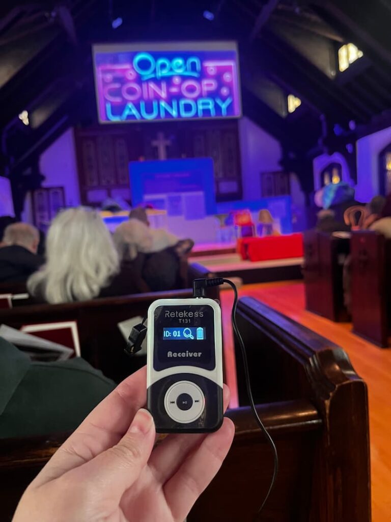 An audience member holds a Retekess T131 receiver, tuned to channel 01, in front of the “Lost Sock Laundry” set — a neon 'Open Coin-Op Laundry' sign glowing above the altar of the church.