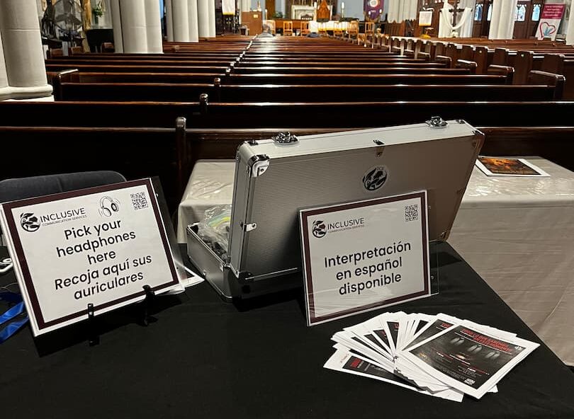 The ICS access table set up at the back of the church before a performance: an open flight case of headsets, a sign reading 'Pick your headphones here / Recoja aquí sus auriculares', a second reading 'Interpretación en español disponible', and a fanned stack of programmes.