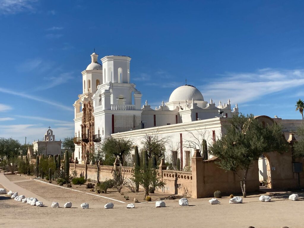 A white Spanish colonial mission church with two towers, one finished with a dome and one left flat-topped, an ornate carved brown facade between them, and a walled courtyard of cacti in front under a clear blue sky.