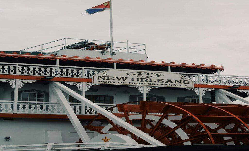 The stern of a paddlewheel riverboat, white gingerbread railings and orange scalloped trim above a red paddlewheel, with a nameboard reading City of New Orleans, Port of New Orleans, LA.