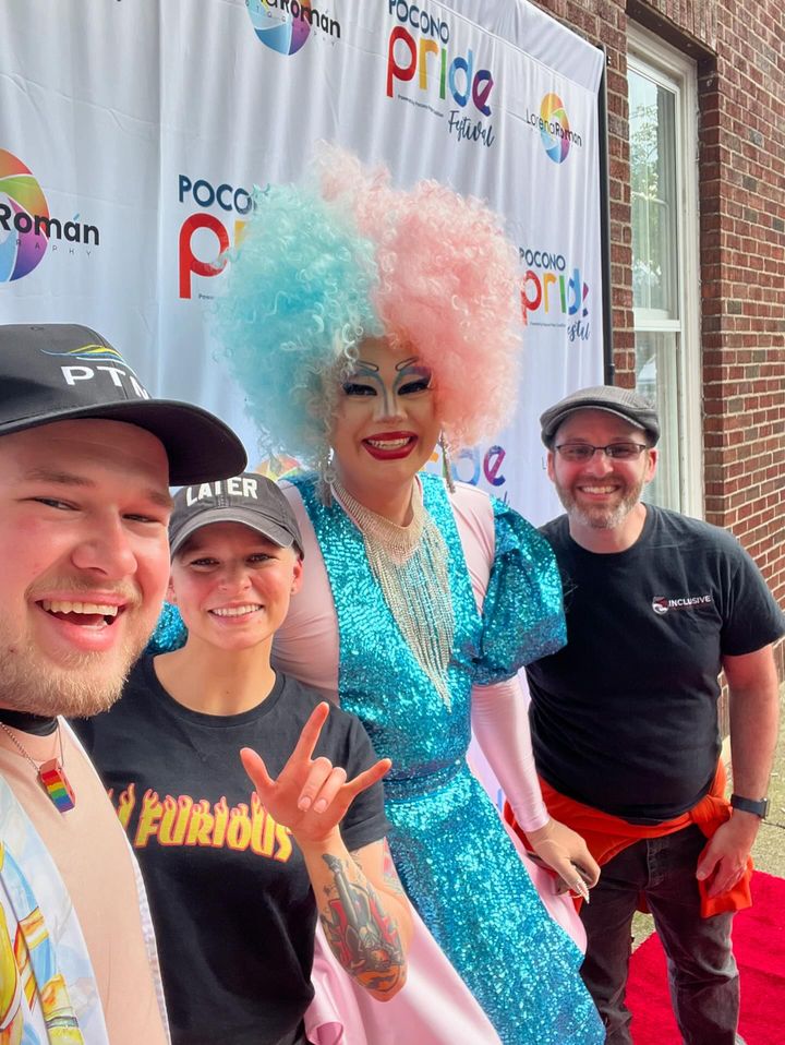 A Joyful group of people poses at the Poconos Pride Festival, with a drag queen in colorful attire centered in front of the pride banner.