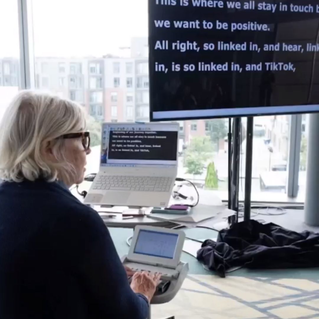A woman types on a stenotype machine as captions appear on a screen behind her.