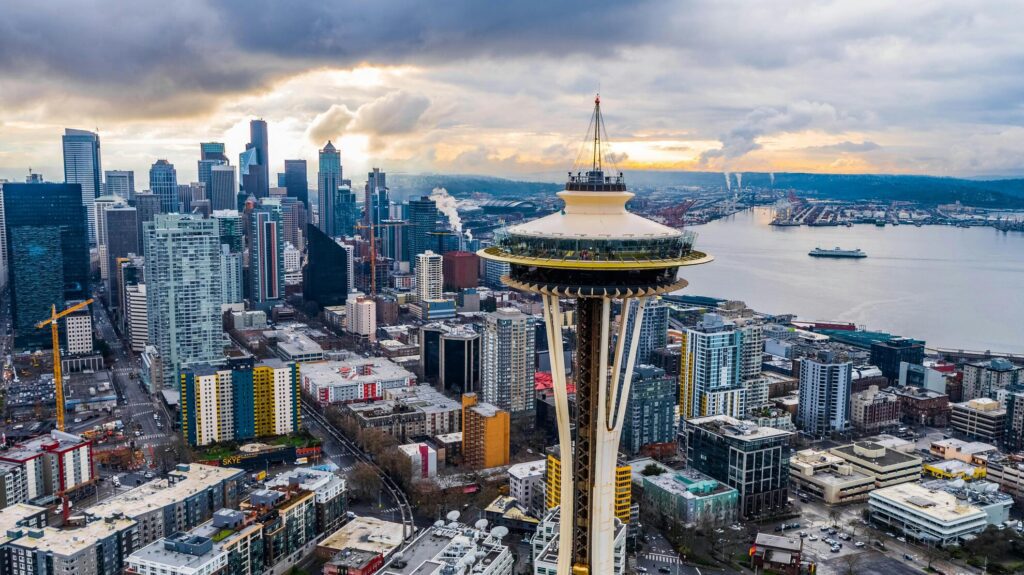 An aerial view of the Space Needle's saucer and tapered legs in the foreground, the downtown Seattle towers behind it to the left and a ferry crossing open water to the right.