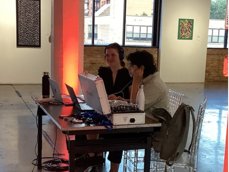 Two interpreters at the interpreting table, one in headphones speaking into a microphone transmitter and the other holding an earbud in place, working from a direct audio feed running into the laptop between them.