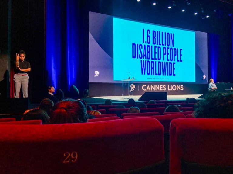 An auditorium with a large screen displaying “1.6 Billion Disabled People Worldwide.” A sign language interpreter stands to the left.