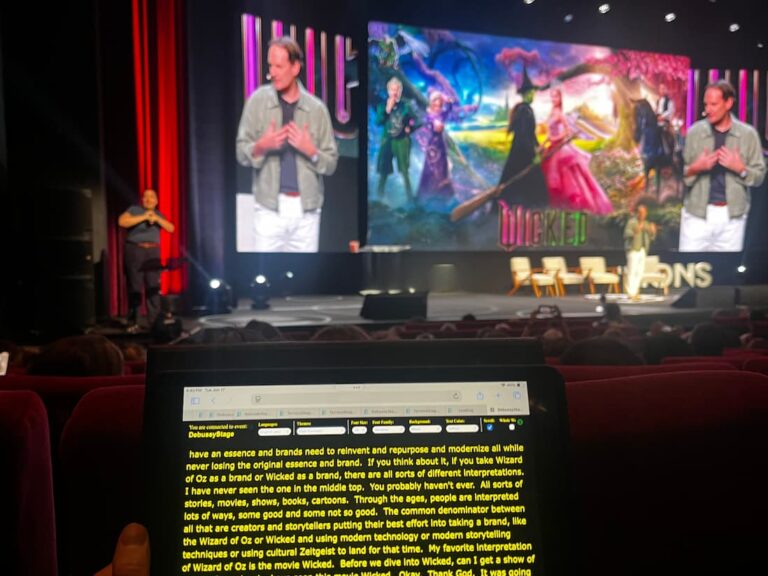 A conference stage with a speaker, a sign language interpreter, and a large projector screen. In the foreground, a tablet displays live captioning.