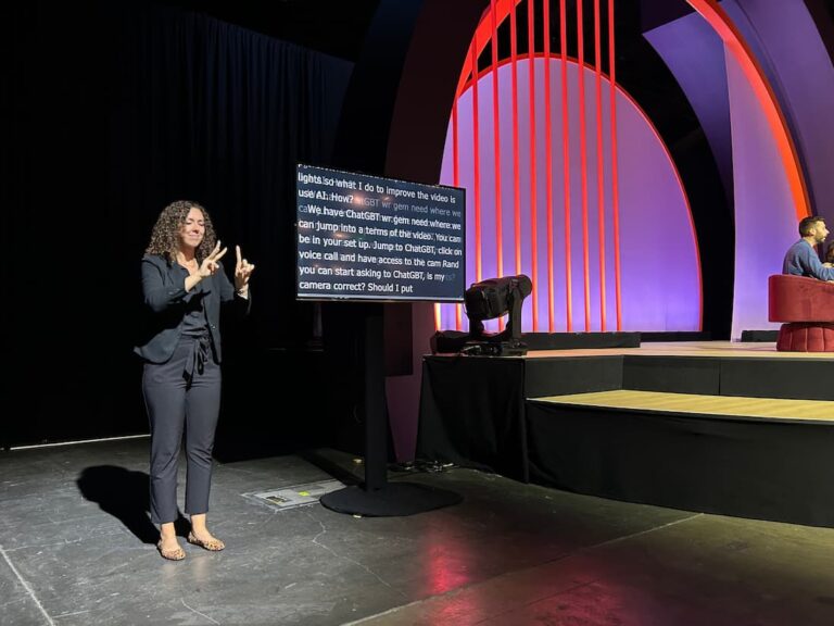 A woman interpreter signs in front of a stage with colorful arches. A large monitor displays the live captioning of the event.