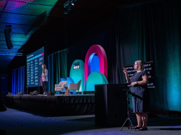 A conference stage with a speaker presenting and a sign language interpreter. A large screen displays a presentation on stage.