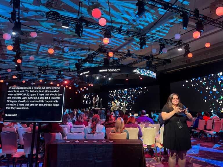 Conference auditorium with multiple large projector screens and colorful lighting. An ASL interpreter stands to the right and a monitor with live captions is displayed on the right.