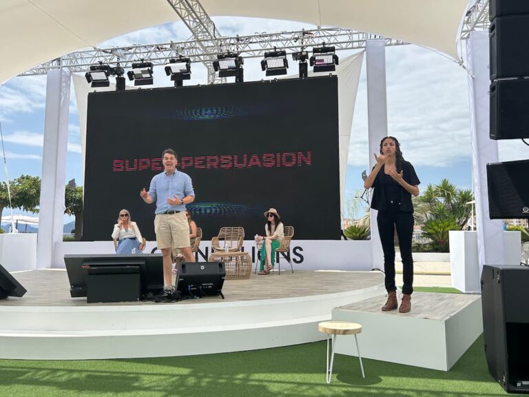 Outdoor stage with speaker in light blue shirt, large screen that says “SUPERPERSUASION,” seated audience, and a sign language interpreter.