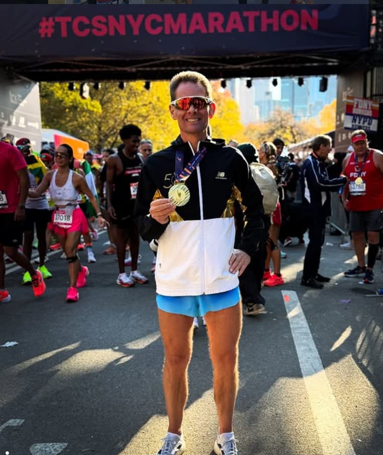 Deaf runner Thomas Eller stands past the finish line under a #TCSNYCMARATHON banner, holding up his marathon finisher medal.