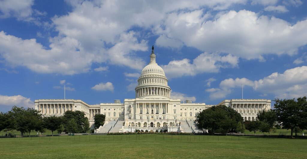 The west front of the United States Capitol under banks of summer cloud, its dome above the terraced steps and the open lawn below.