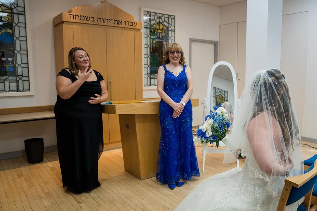 In a church, a bride in a white wedding gown and veil is seated, holding a bouquet of white and blue flowers, while facing two women. One woman, dressed in a black gown, is signing in ASL, and the other, in a blue sequined dress, smiles warmly.