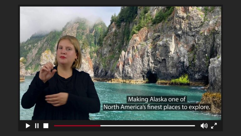 A travel tour video showing the rugged, steep cliffs and vibrant turquoise waters of Alaska's coastline. This video includes captions and a sign language interpreter, dressed in black.