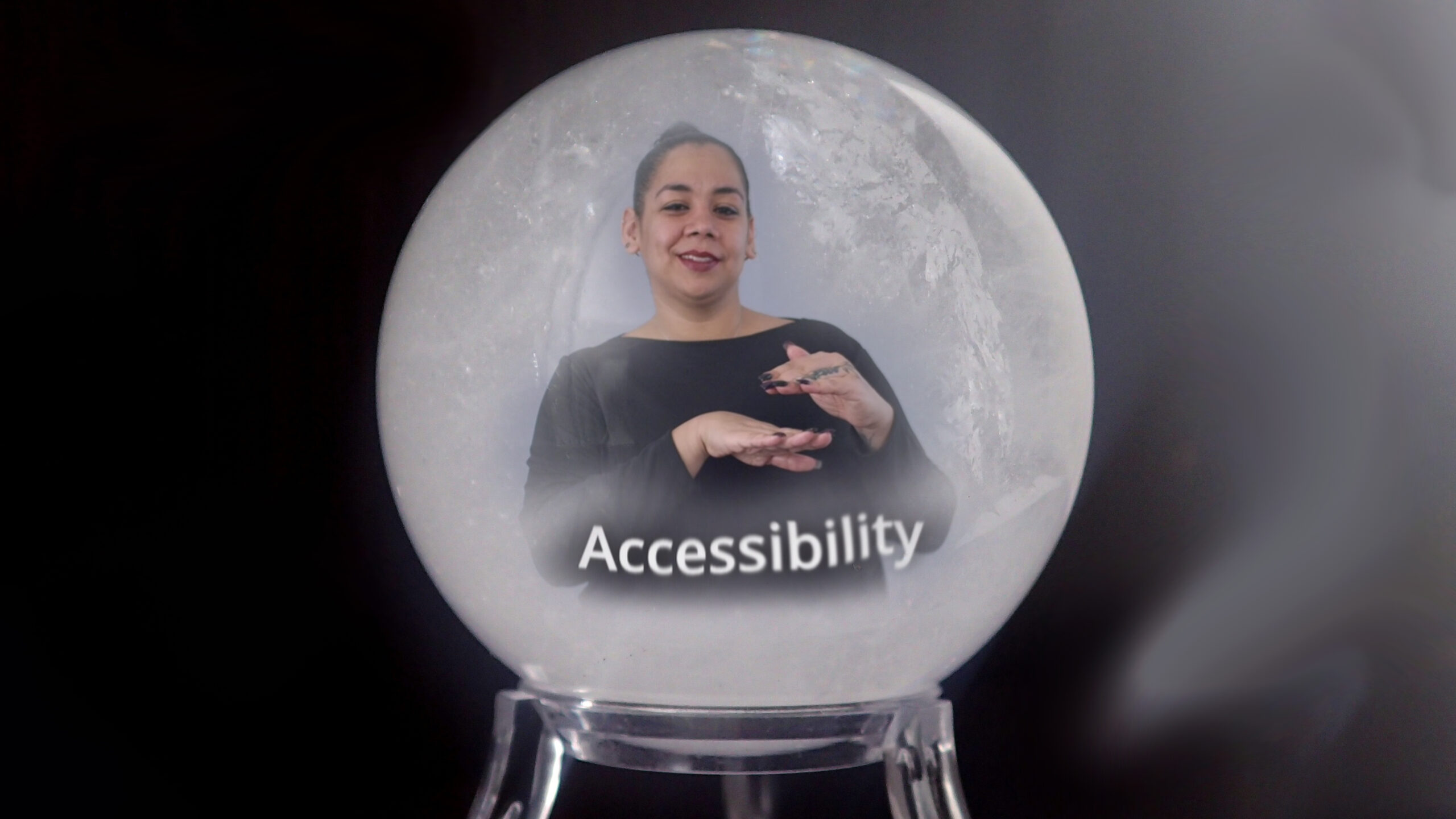 The image showcases a snow globe effect featuring a woman who appears to be signing, indicating the concept of "Accessibility." The woman is situated in the center, dressed in a dark outfit, with her left hand raised and her right hand positioned below it, creating a gesture that likely represents communication or signing. The background shows a softly blurred snowy texture, enhancing the wintery aesthetic of a snow globe. The word "Accessibility" is prominently displayed at the bottom of the globe in white text, making it a focal point. The overall color scheme combines dark tones with lighter shades creating a contrast that emphasizes the subject matter.